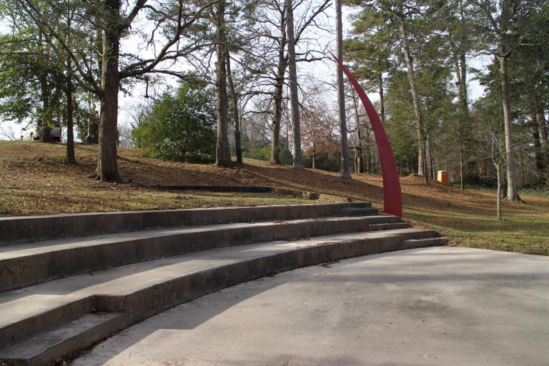 Image of the outside cement stairs with the Blade sculpture at the end. It is a tall, red, four-sided base that comes to a sharp point at the top, curving slightly.